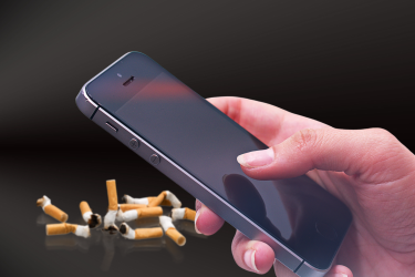 A hand holding a mobile phone with crushed cigarettes in the background