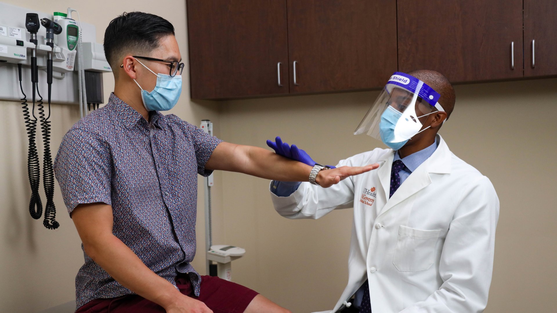 A shot of a patient and a doctor in PPE gear representing COVID time. The doctor is examining the patient sitting in the clinic room.