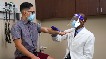 A shot of a patient and a doctor in PPE gear representing COVID time. The doctor is examining the patient sitting in the clinic room.