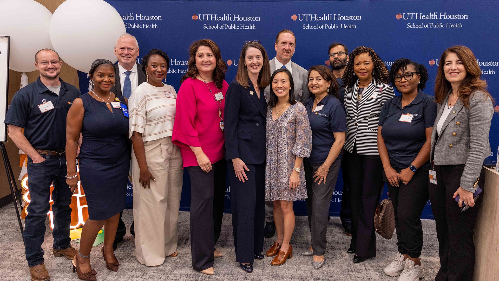 Group of staff and attendees pose for a photo in front of a UTHealth Houston School of Public Health backdrop at an indoor event.