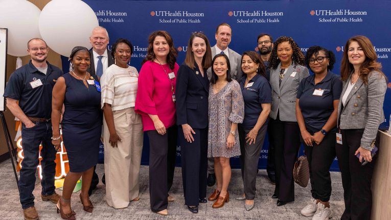 Group of staff and attendees pose for a photo in front of a UTHealth Houston School of Public Health backdrop at an indoor event.
