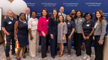 Group of staff and attendees pose for a photo in front of a UTHealth Houston School of Public Health backdrop at an indoor event.