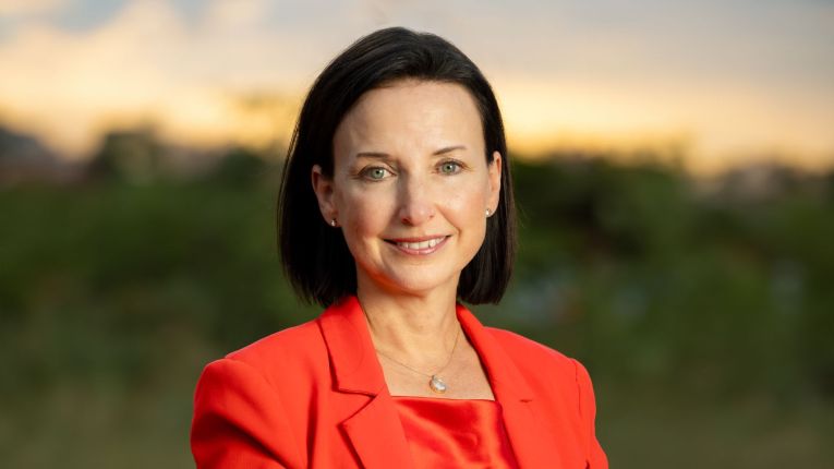 A professional headshot of a woman with short dark hair and green eyes, wearing a bright red blazer and a delicate necklace, smiling at the camera. The background is softly blurred with greenery and a warm sunset sky.