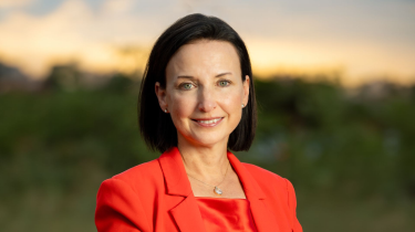 A professional headshot of a woman with short dark hair and green eyes, wearing a bright red blazer and a delicate necklace, smiling at the camera. The background is softly blurred with greenery and a warm sunset sky.