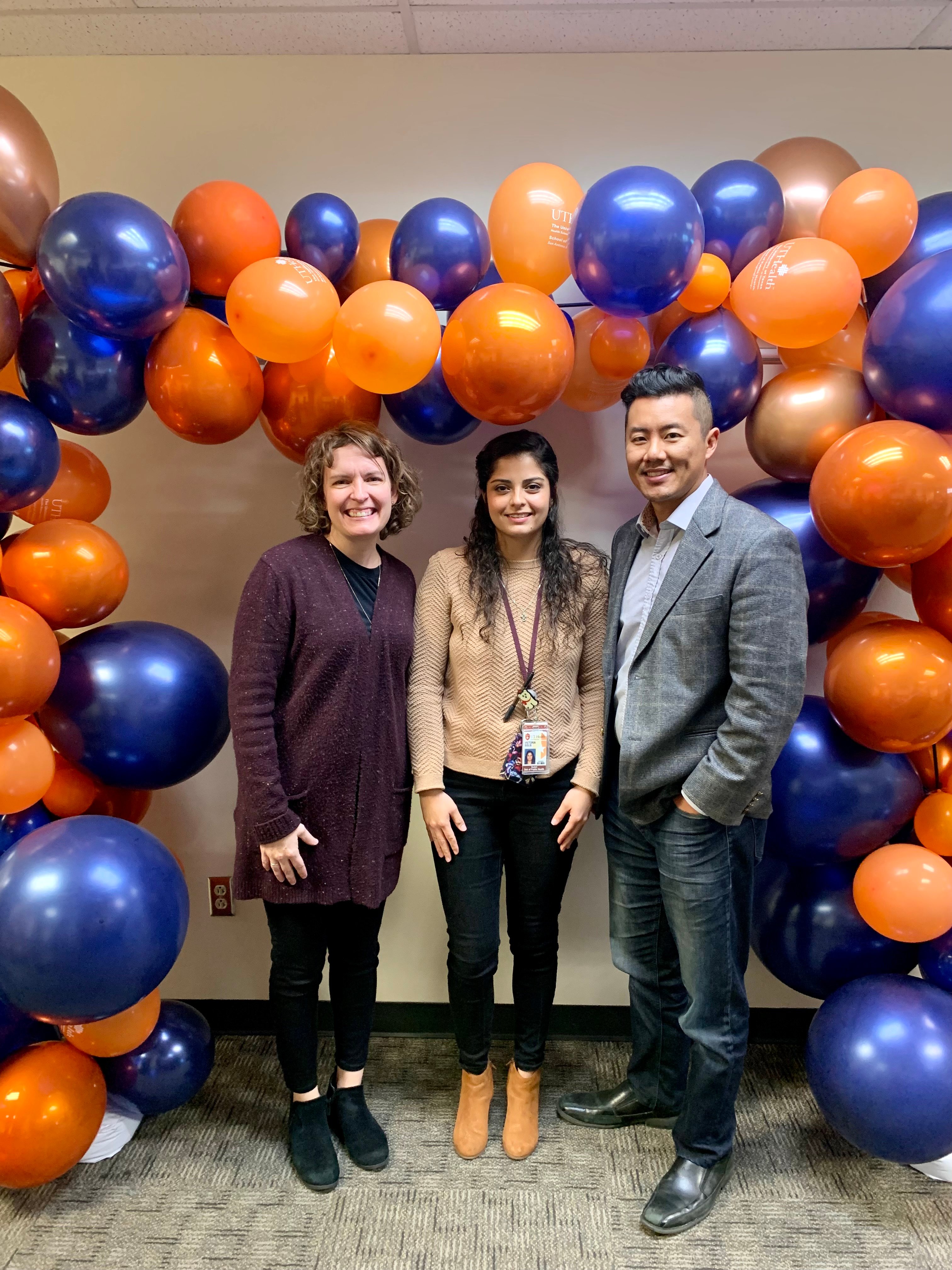 Victoria Solis stands between a man and a woman under an arch of orange and blue balloons