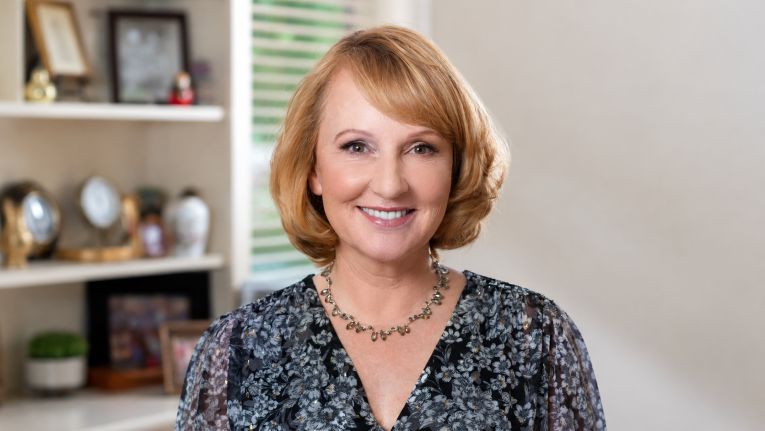 Smiling woman with short blonde hair and blue eyes, wearing a floral blouse and necklace, posed in a softly lit home interior with blurred shelves and framed photos in the background.
