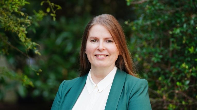 Woman wearing an emerald sport coat and white shirt smiling at camera, standing in front of trees and foliage.