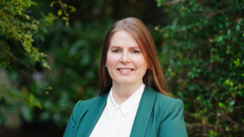Woman wearing an emerald sport coat and white shirt smiling at camera, standing in front of trees and foliage.