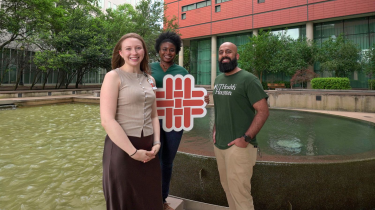 Three students standing in front of fountain holding a school logo sign.