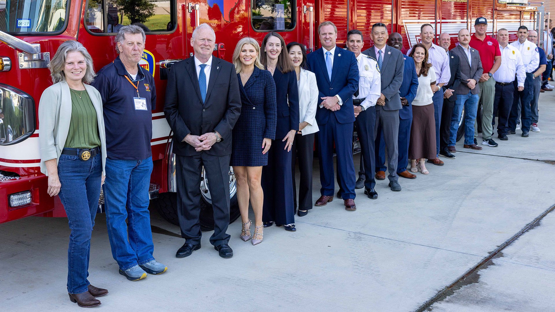 Group of community leaders, firefighters, and officials standing in a line beside several red fire trucks at a fire station.